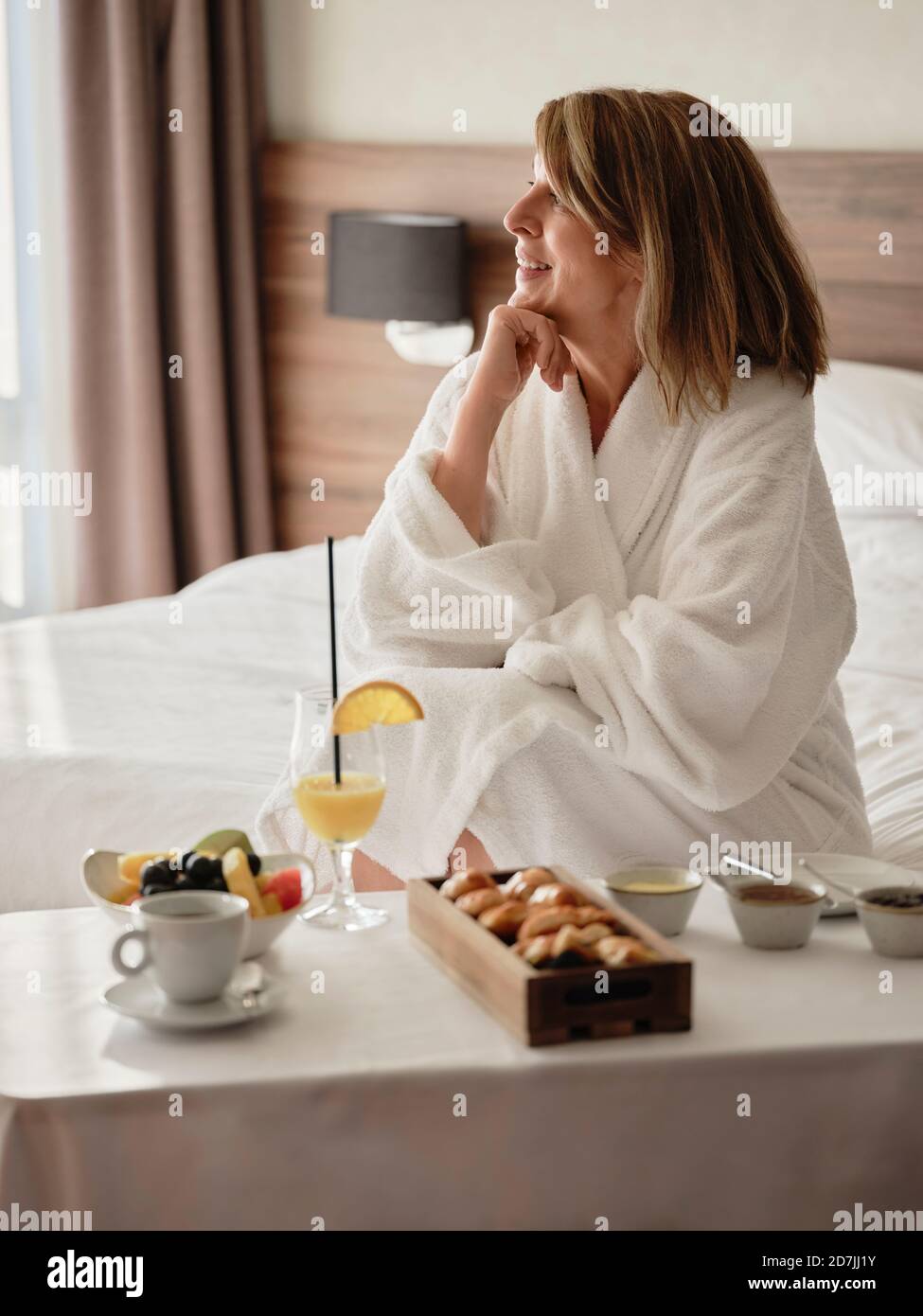 Lächelnde, nachdenkliche ältere Frau, die beim Frühstück wegschaute In einem luxuriösen Hotelzimmer Stockfoto