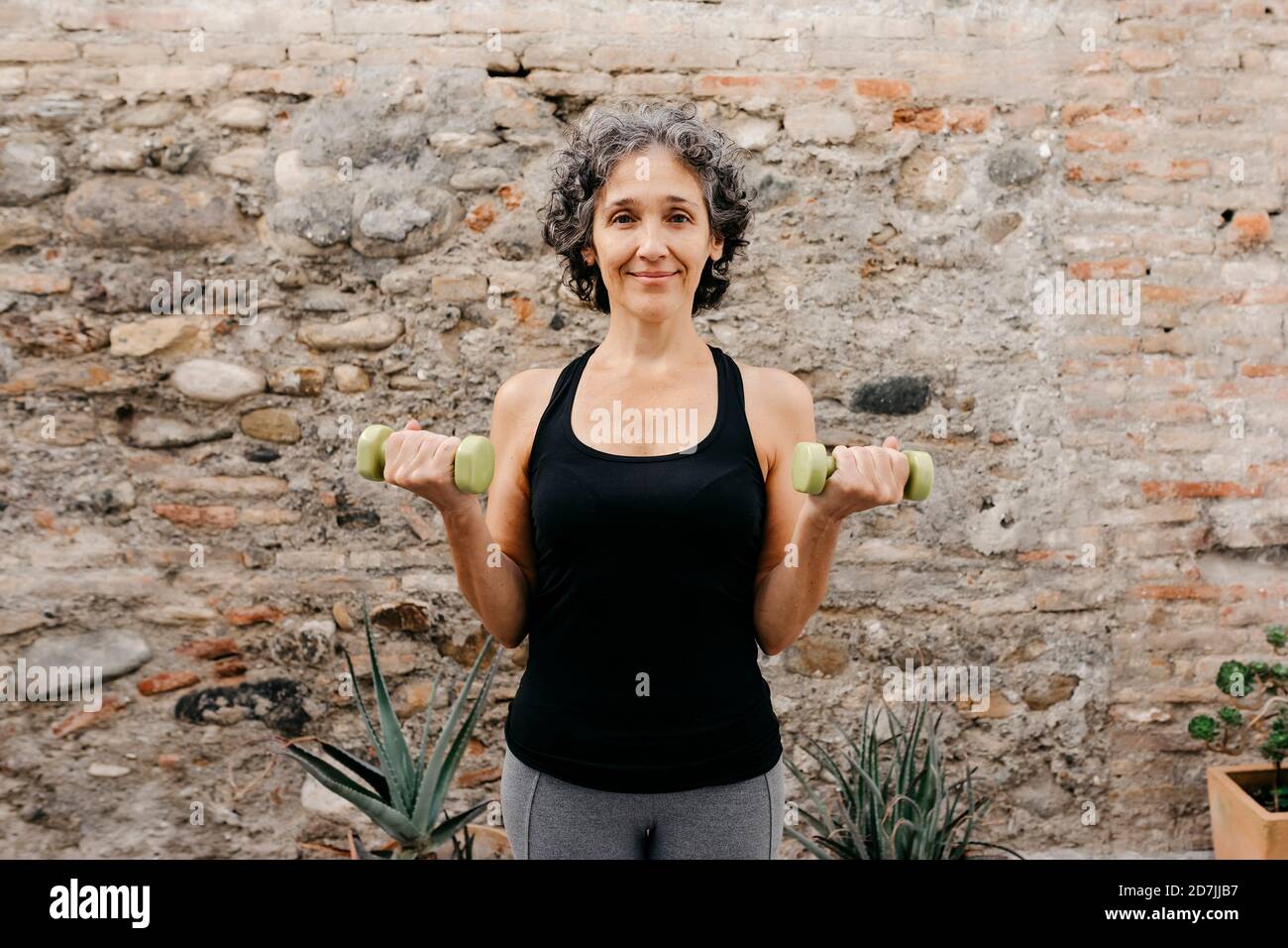 Lächelnde Frau, die mit Hanteln gegen die Steinwand am Rücken trainiert Hof Stockfoto