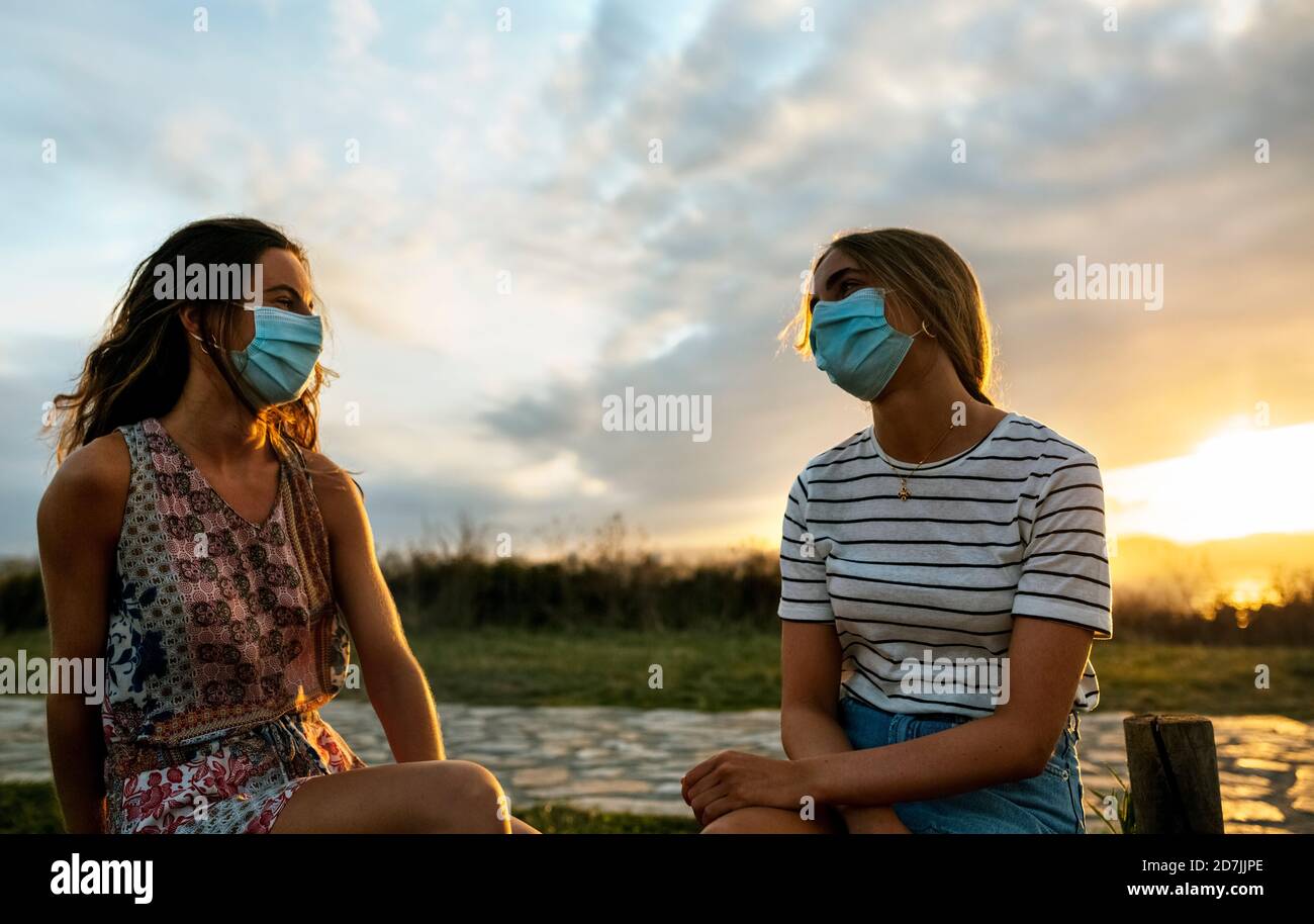 Frauen in schützenden Gesichtsmasken halten soziale Distanz während des Sitzens Gegen den Himmel bei Sonnenuntergang Stockfoto