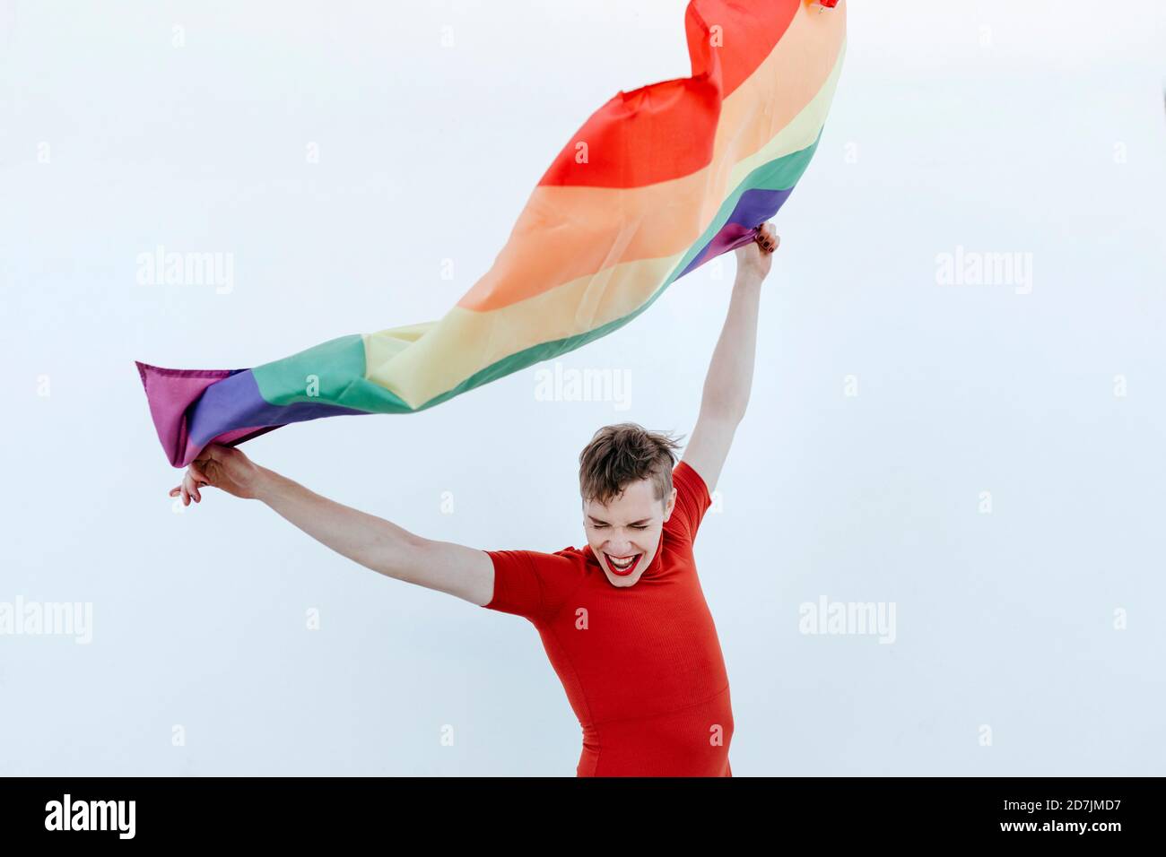 Nicht-binäre Person winkt mehrfarbige Flagge, während sie gegen Weiß steht Wand Stockfoto