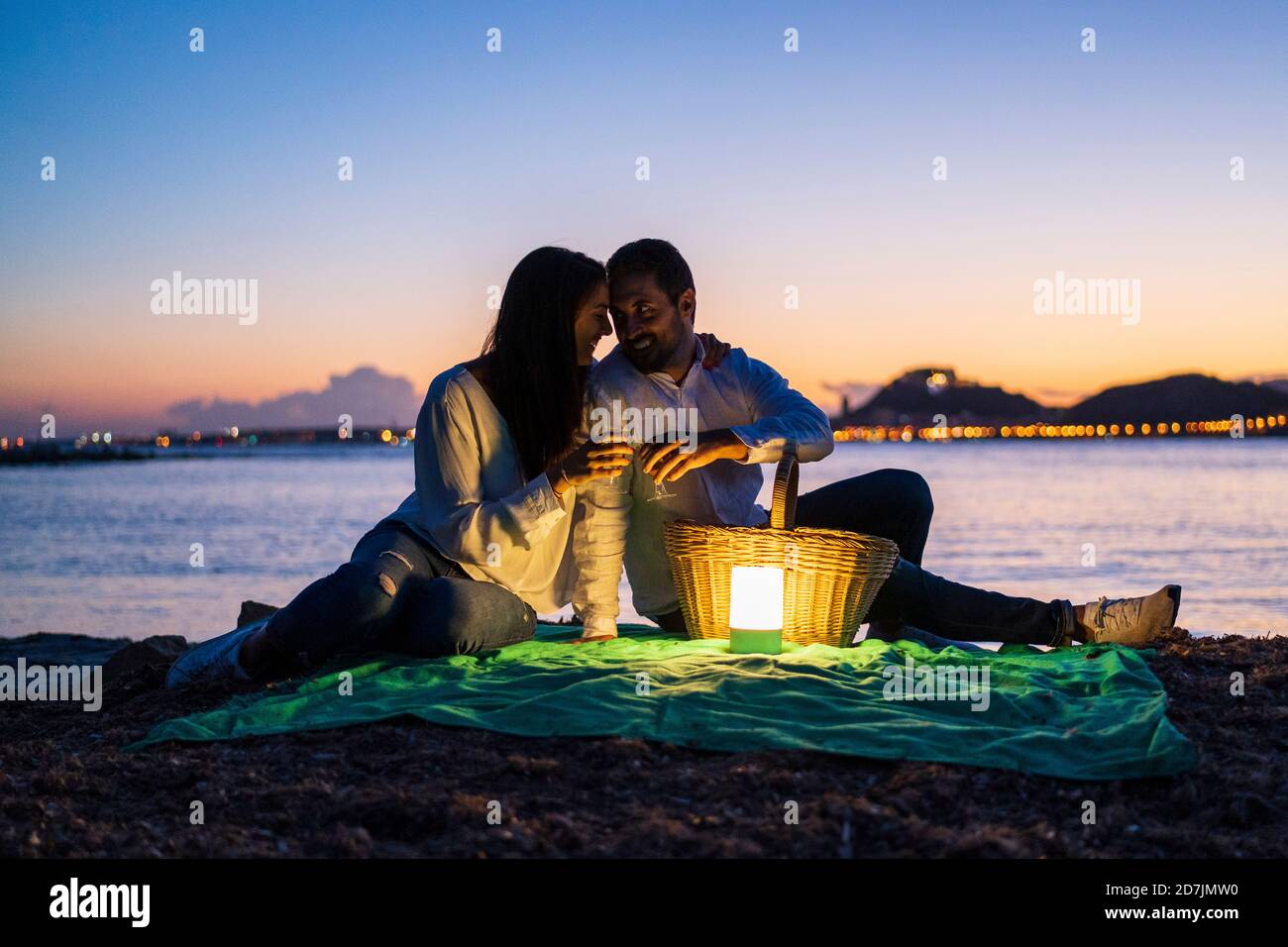 Romantisches Paar mit Getränken am Strand gegen klaren Himmel sitzen Während der Dämmerung Stockfoto