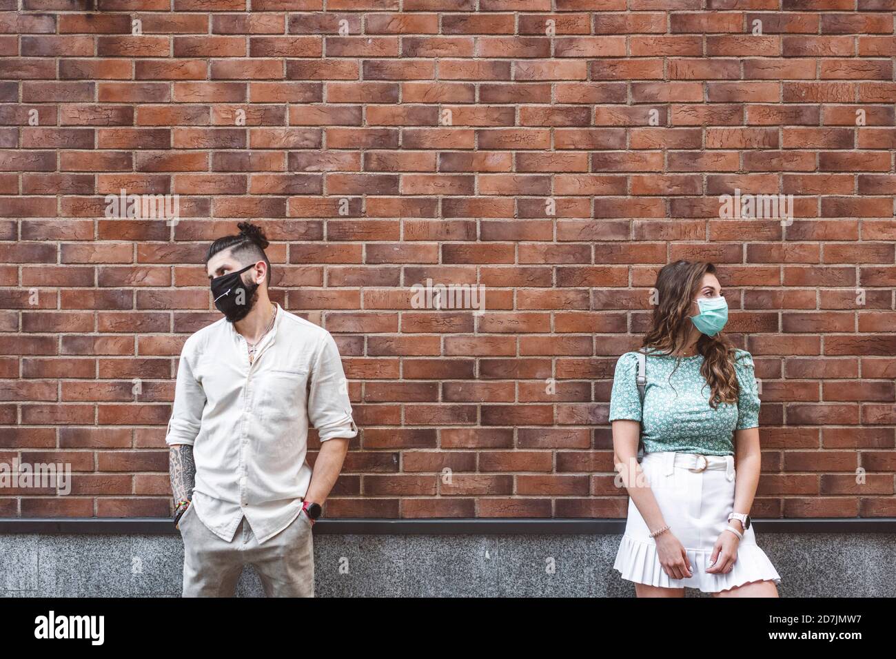 Trauriges Paar in schützenden Gesichtsmasken, die gegen eine Ziegelwand stehen Während der Coronavirus-Krise Stockfoto