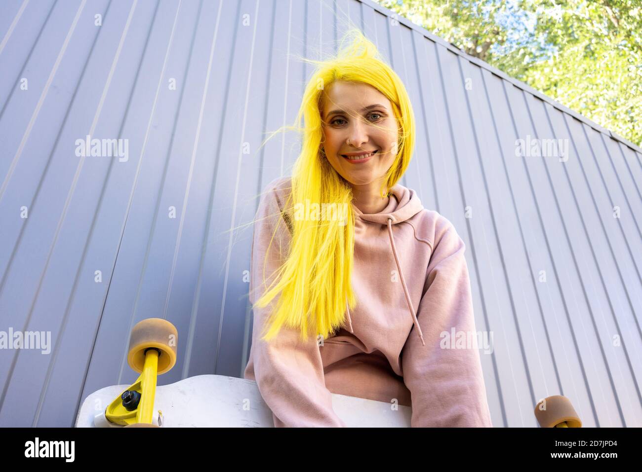 Lächelnd gefärbte gelbe Haare Frau mit Skateboard gegen Metall stehen Wand Stockfoto