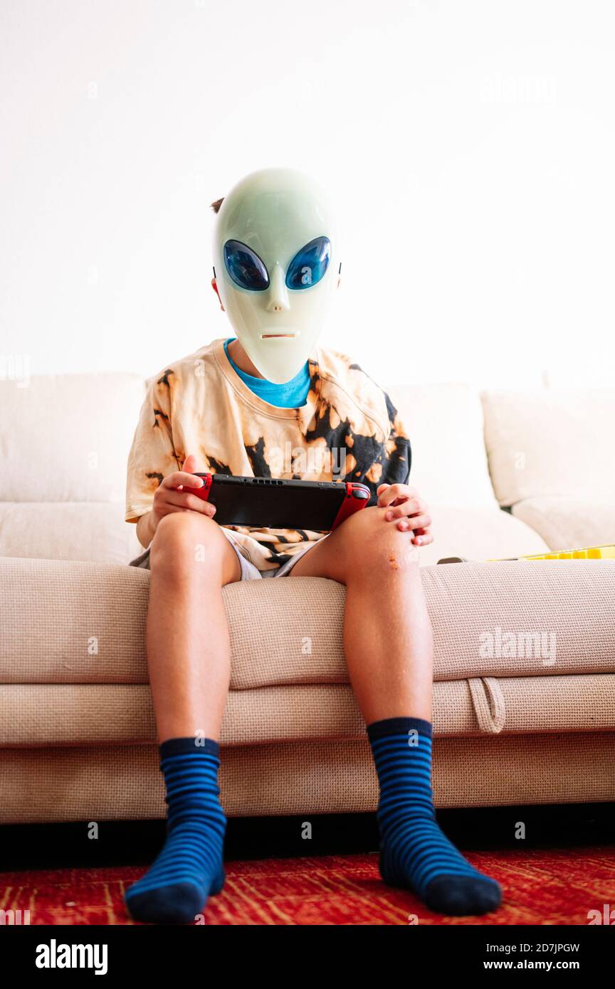 Junge trägt Alien Maske spielen Handheld-Videospiel während des Sitzens Auf dem Sofa zu Hause Stockfoto