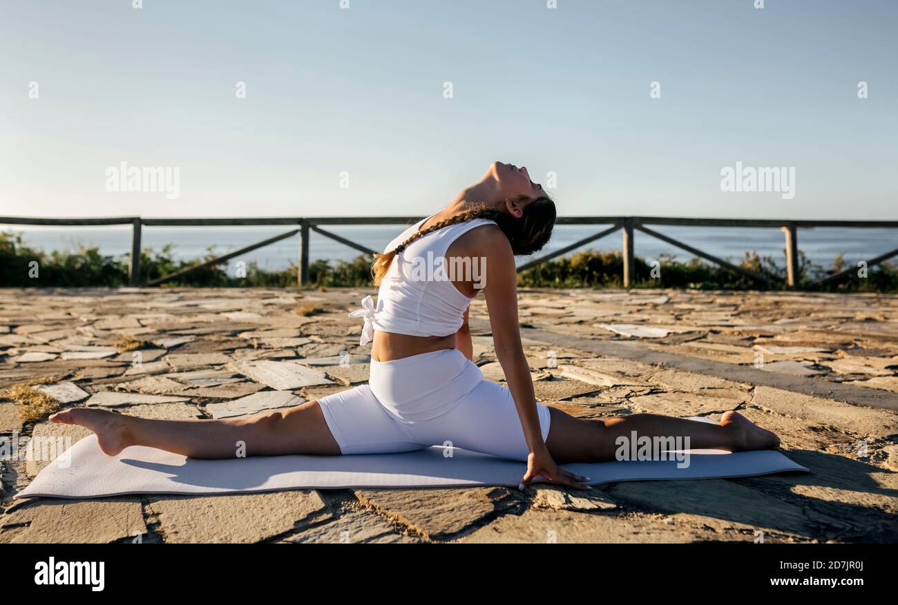 Junge Frau macht Splits auf Trainingsmatte gegen klaren Himmel Bei Sonnenuntergang Stockfoto