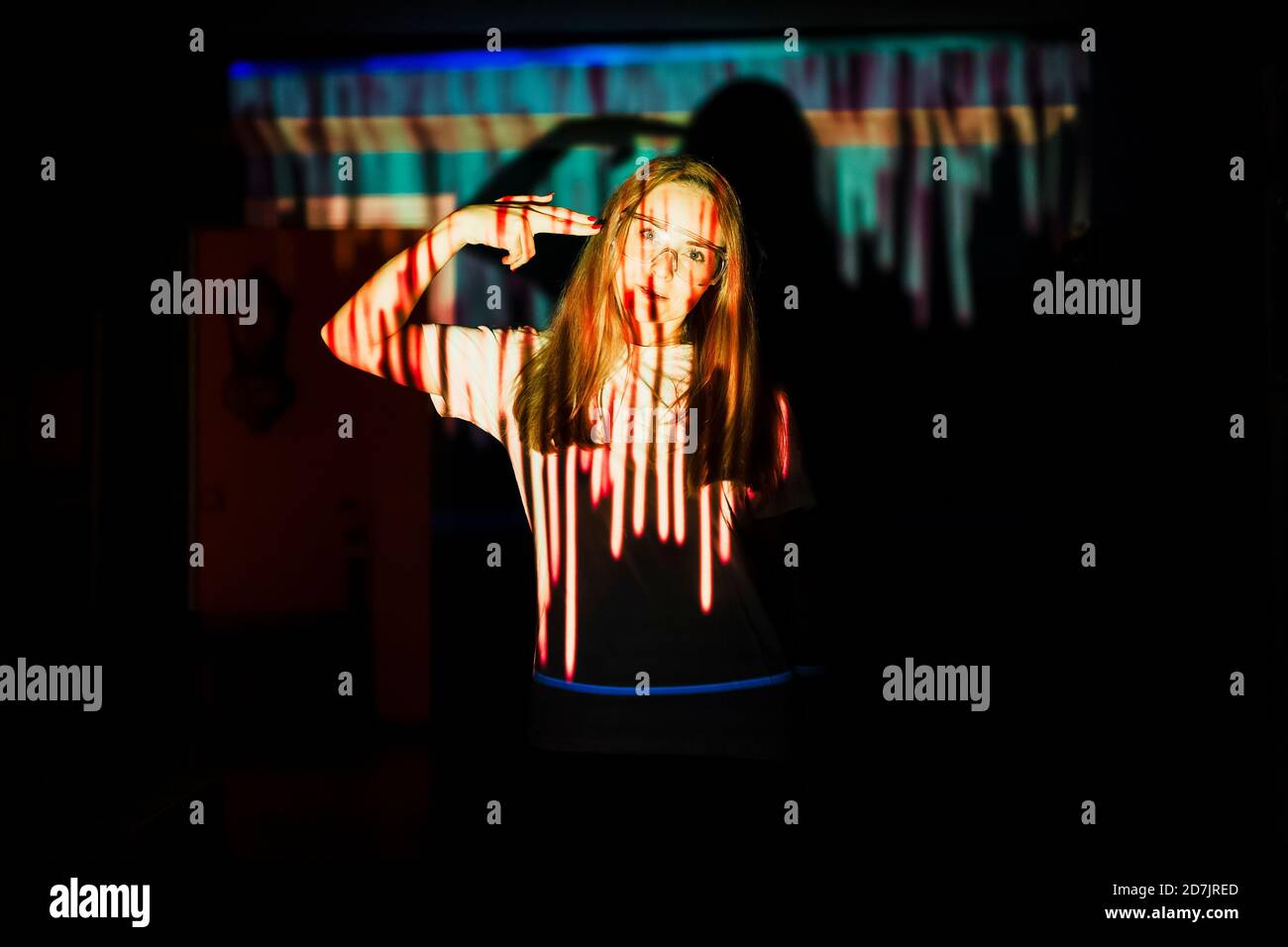 Frau zeigt Pistole Hand Zeichen auf dem Kopf, während im Stehen Lichtreflexion zu Hause während einer Pandemie Stockfoto