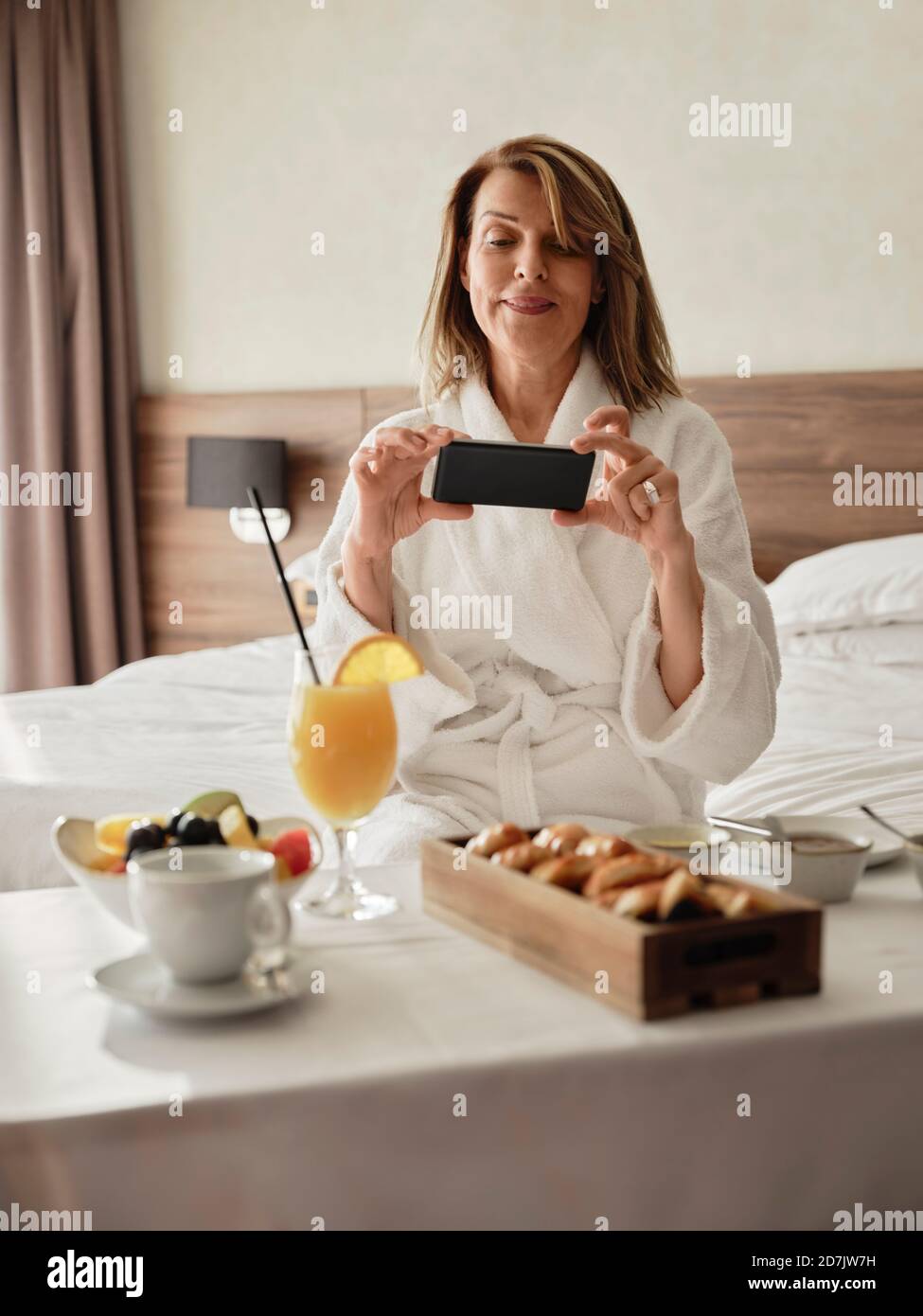 Lächelnde blonde ältere Frau fotografiert Frühstück über Smartphone während Sitzen auf dem Bett im Hotelzimmer Stockfoto