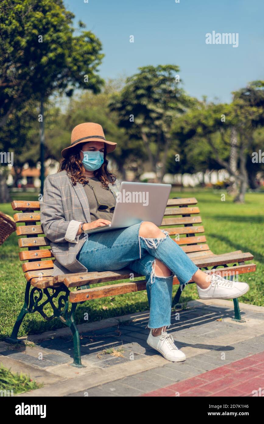 Vertikales Bild einer Bloggerin, die im Freien auf ihrem Laptop tippt, während sie eine medizinische Maske trägt und ein Fahrrad fährt. Neues normales Konzept. Speicherplatz kopieren Stockfoto