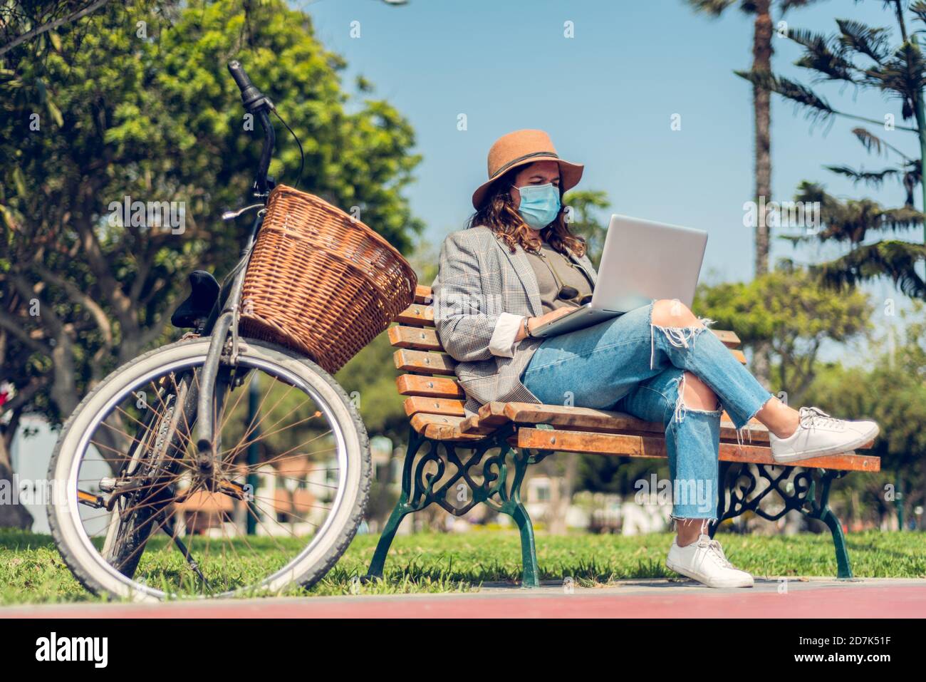 Frau arbeitet in einem Park sitzend und schützt sich mit einer medizinischen Maske. Porträt einer modischen Frau, die die Natur genießt und schreibt auf ihrem Blog Stockfoto