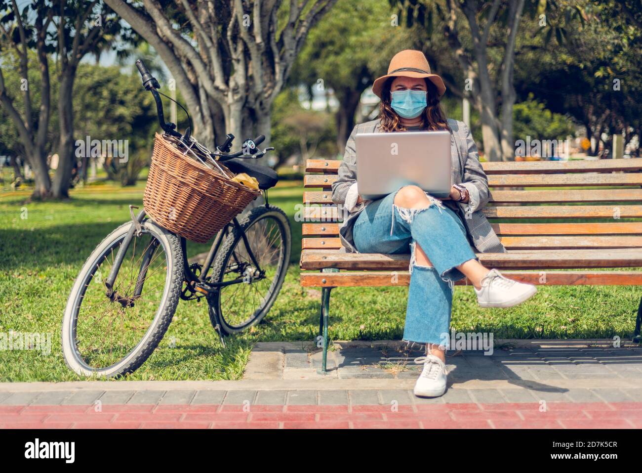 Frau arbeitet in einem Park sitzend und schützt sich mit einer medizinischen Maske. Porträt einer modischen Frau, die die Natur genießt und schreibt auf ihrem Blog Stockfoto
