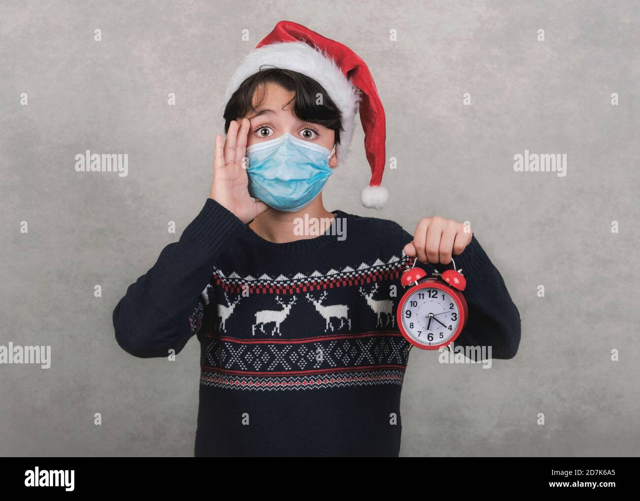 Frohe Weihnachten, Lustiges Kind mit medizinischer Maske und Wecker trägt Weihnachtsmann Hut auf grauem Hintergrund Stockfoto