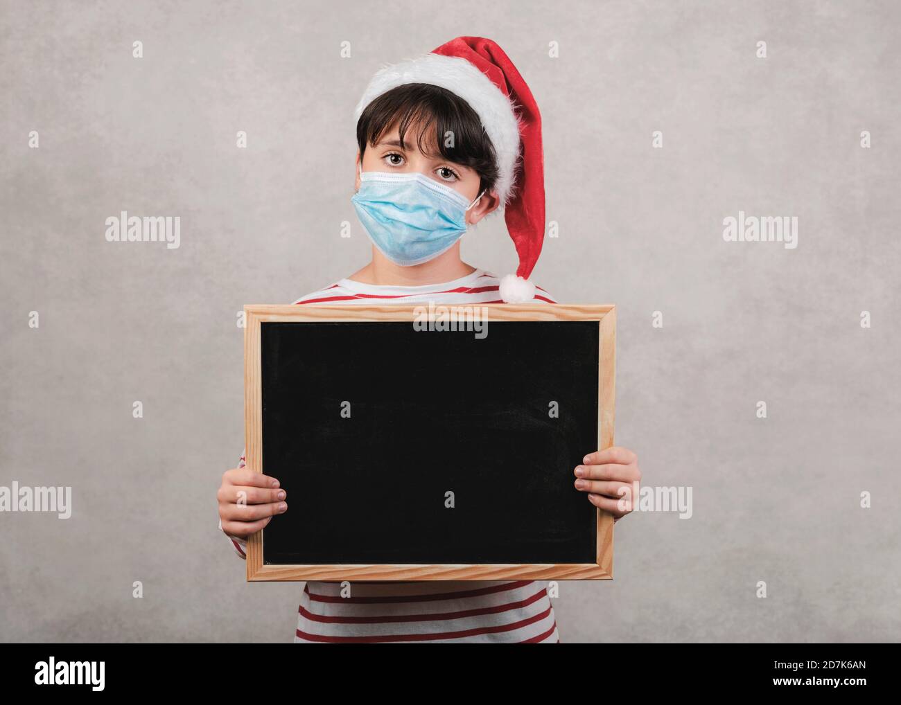 Frohe Weihnachten, lustiges Kind mit medizinischer Maske, die eine Tafel hält Grauer Hintergrund Stockfoto