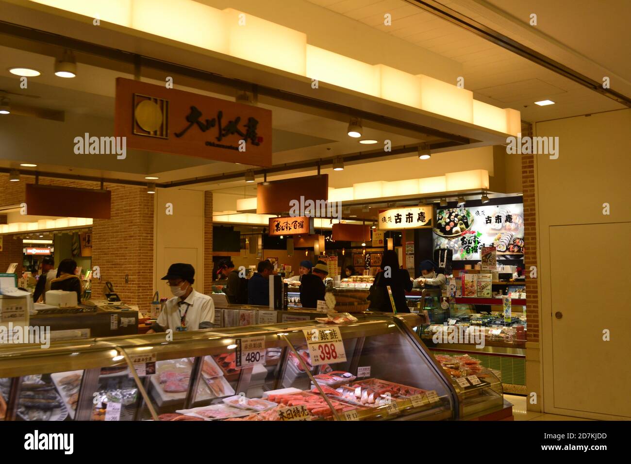 Tokio, Japan-2/27/16: Arbeiter maskiert vor einem Tokioter Fleischmarkt/Deli; hinter ihnen sind Käufer, die ein Geschäft verkaufen, das Bento-Boxen verkauft. Stockfoto