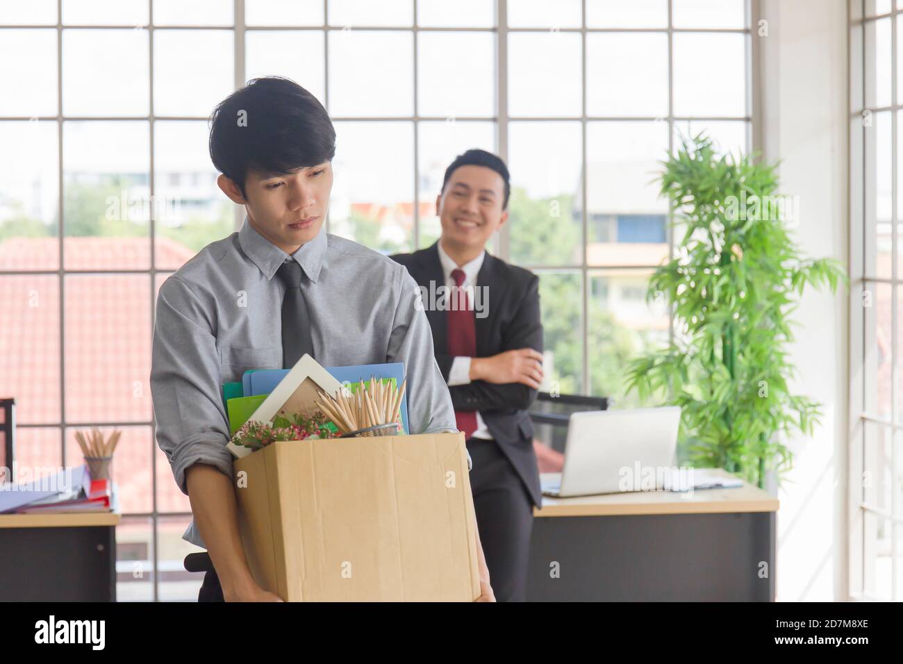 Ein asiatischer Mann, der traurigerweise einen Karton mit seinen persönlichen Sachen in der Hand hält, und sein Manager lächelt hinter ihm auf einem Schreibtisch in einem Büro. Stockfoto