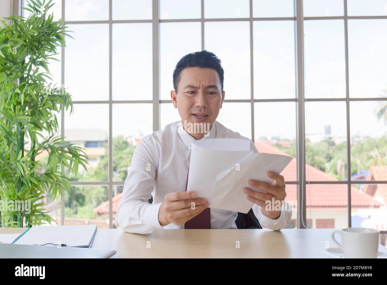 Ein Mann in einer asiatischen Firma erhält mit schockierter Äußerung wichtige Dokumente auf seinem Schreibtisch. Stockfoto