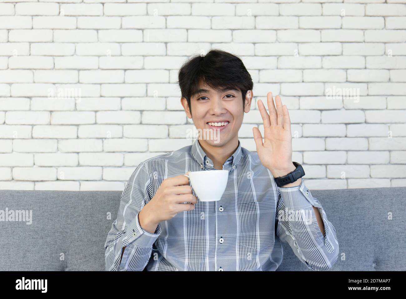 Ein junger asiatischer Mann, hält eine Kaffeetasse und lächelt hell winkend seine Hand. Stockfoto
