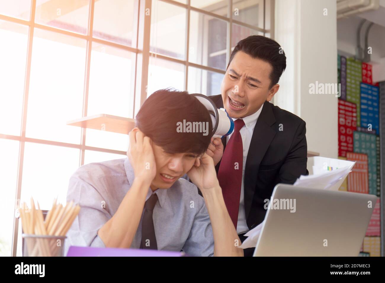 Ein wütender asiatischer Mann-Manager, der in einem Lautsprecher im Ohr seines Untergebenen schreit, um Dokumente zu greifen. Stockfoto