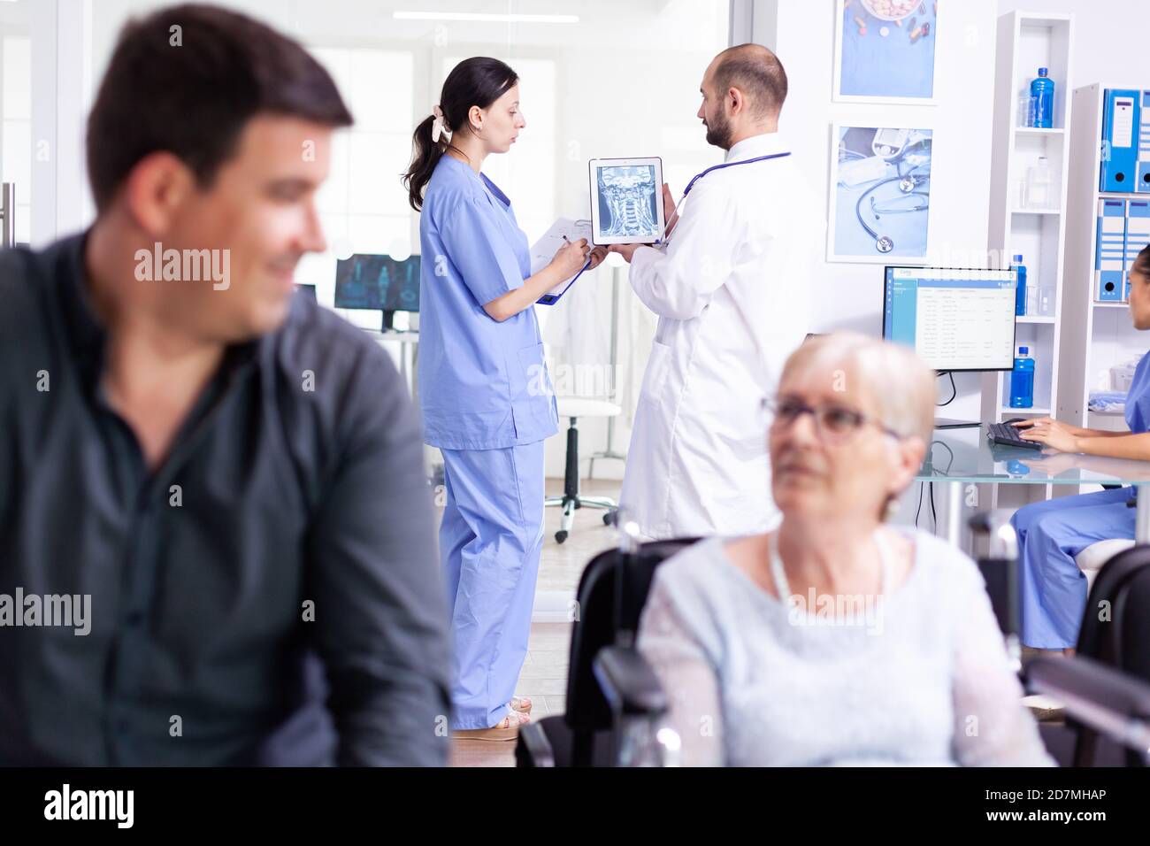 Medizinisches Personal, das einen Tablet-pc mit Radiographie im Krankenhausflur hält. Arzt, der die Diagnose mit der Krankenschwester im Wartebereich der Klinik bespricht. Alte Frau sitzt im Rollstuhl. Stockfoto