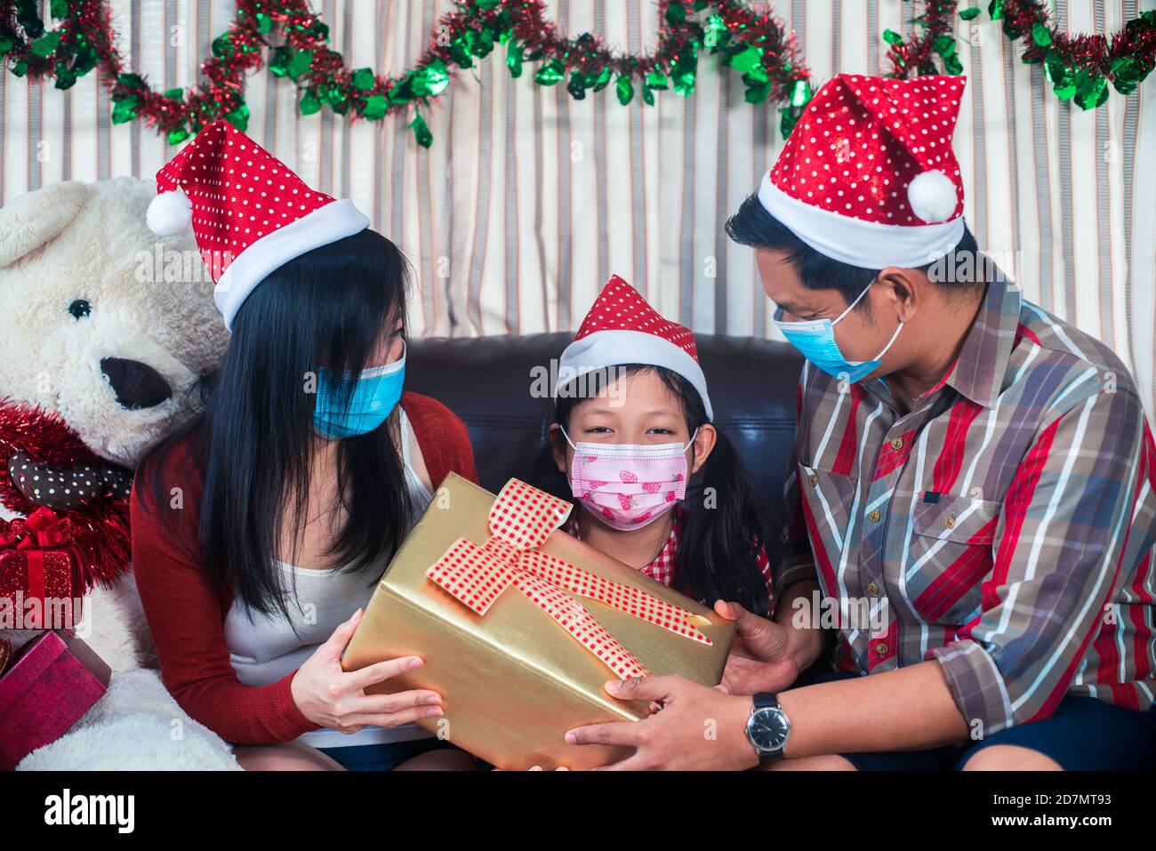 Glückliche Eltern geben Weihnachten Geschenk für Tochter mit tragen Gesicht Maske.Familie im weihnachtlichen Interieur Stockfoto