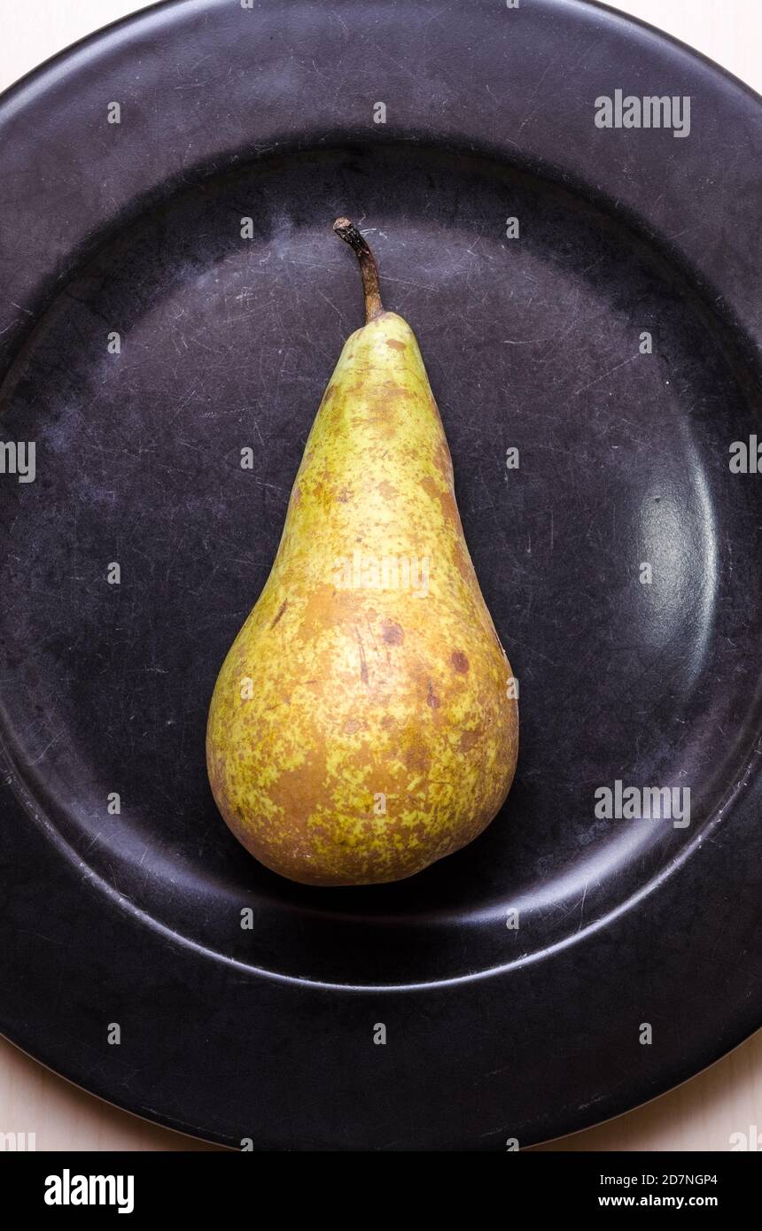 Eine einzelne Birnenfrucht, Pyrus, flach auf dunkelschwarzem Rundteller auf Holztisch oder Schreibtisch, Food-Fotografie, Nahaufnahme, Kopierraum Stockfoto