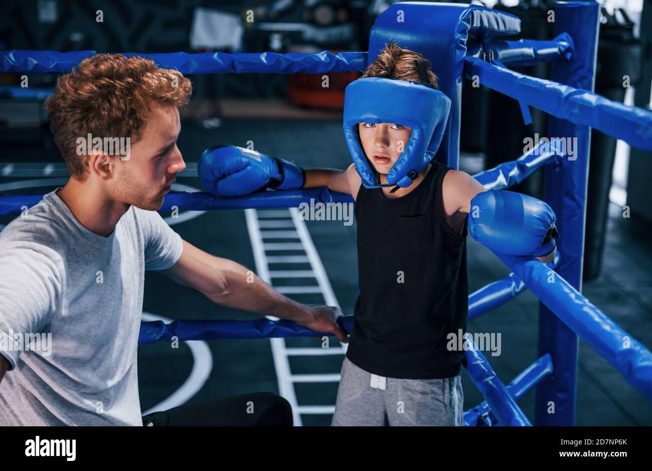 Junge Boxtrainer hilft kleinen Jungen in Schutzkleidung Auf dem Ring zwischen den Runden Stockfoto