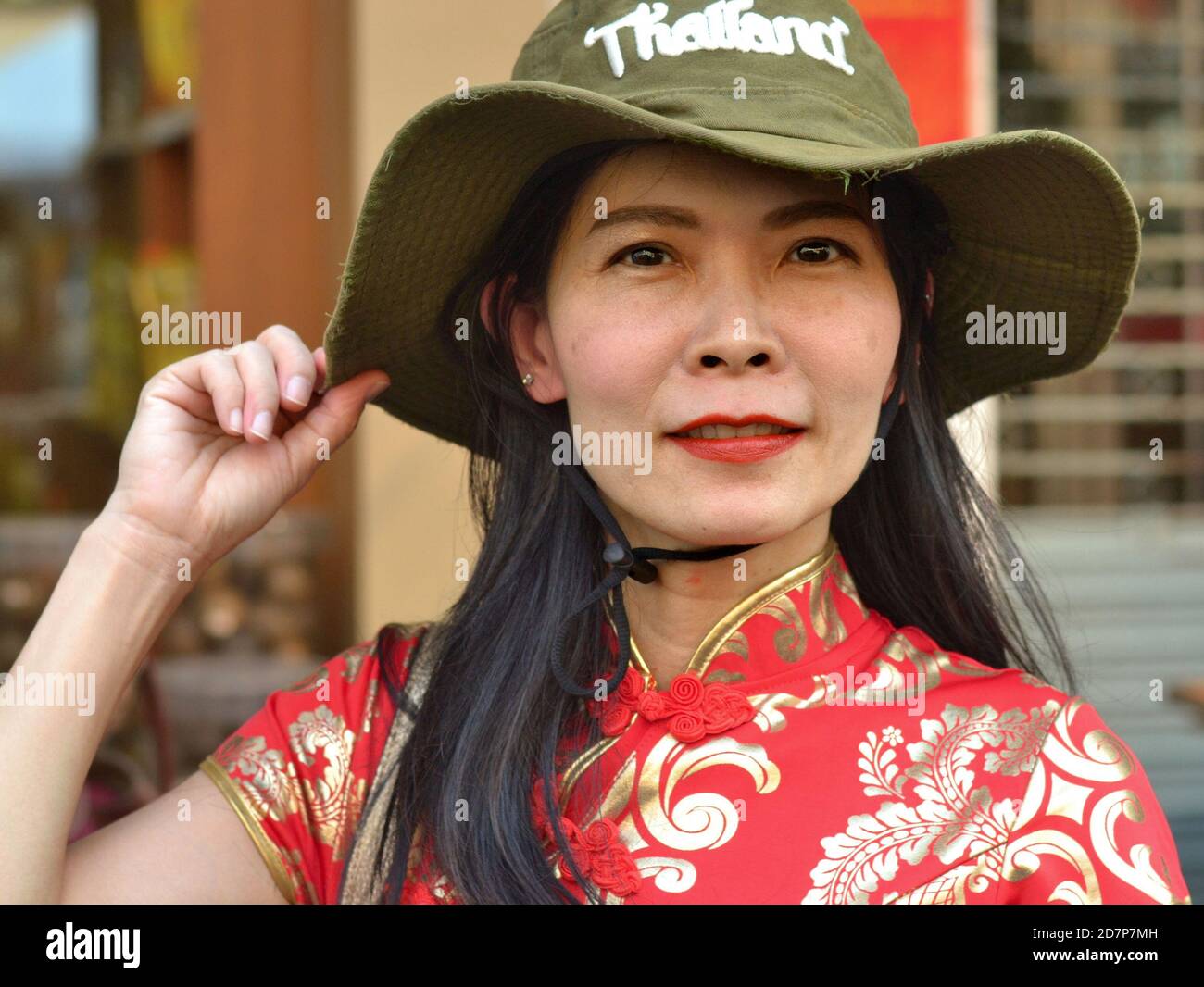 Die Thailändische Chinesin mittleren Alters trägt ein traditionelles chinesisches Kleid in Rot und Gold (Cheongsam) und einen grünen, breitkrempigen Thailand-Souvenir-Buschhut. Stockfoto