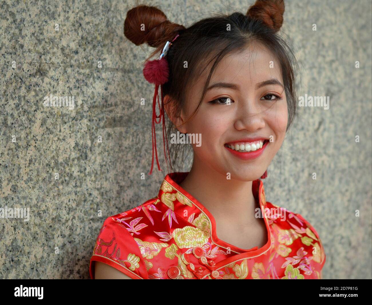 Junge Thai-chinesische Schönheit mit traditionellen chinesischen Doppelseitenbrötchen trägt ein rot-goldenes chinesisches Seidenkleid (Cheongsam) und lächelt für die Kamera. Stockfoto