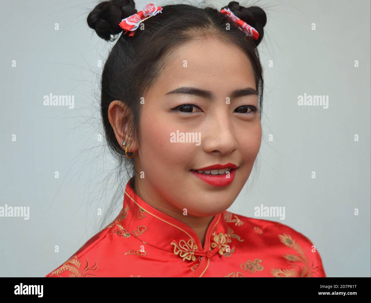 Junge Thailändische Chinesin mit trendigen Kontaktlinsen und traditionellen chinesischen Doppelseitenbrötchen trägt ein rotes chinesisches Kleid und lächelt für die Kamera. Stockfoto