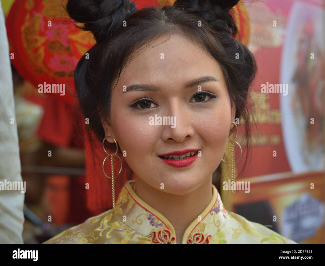 Junge Thai chinesische Schönheit mit trendigen Kontaktlinsen und traditionellen Doppel-Seite Brötchen trägt ein klassisches chinesisches Seidenkleid und lächelt für die Kamera. Stockfoto