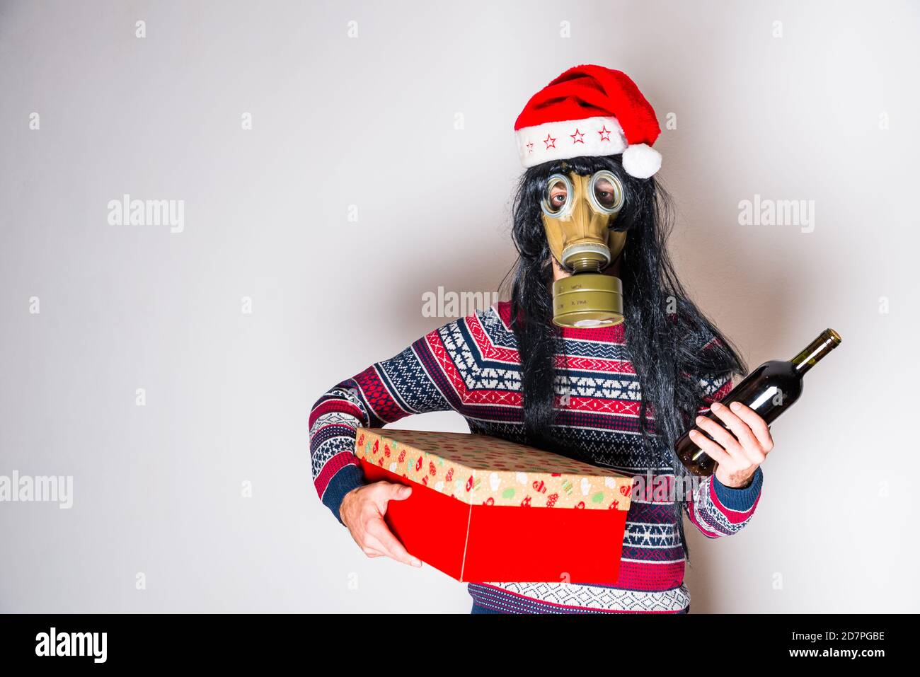 Person mit langen schwarzen Haaren in Gasmaske und weihnachten hut mit Geschenk und Flasche Wein in Händen Heller Hintergrund mit Kopierbereich Stockfoto