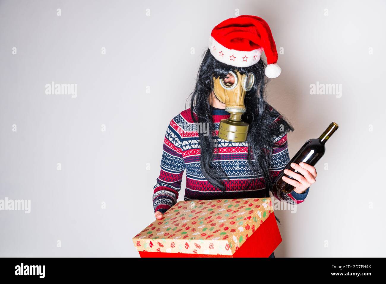 Person mit langen schwarzen Haaren in Gasmaske und weihnachten hut mit Geschenk und Flasche Wein in Händen Heller Hintergrund mit Kopierbereich Stockfoto
