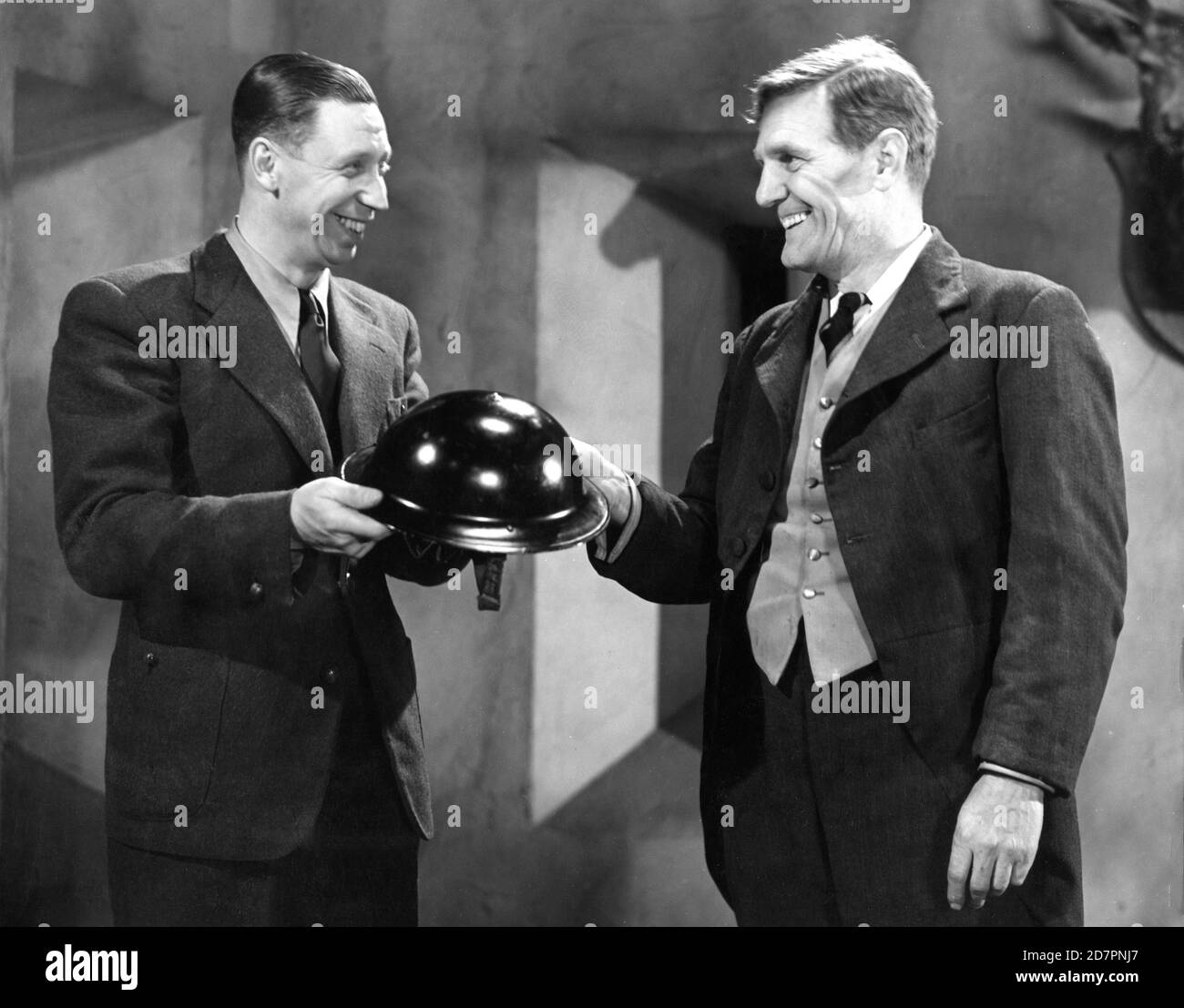 GEORGE FORMBY präsentiert mit Air RAID Helm von WILL HAY am Set Candid während des letzten Tages der Dreharbeiten VON THE GHOST OF ST. MICHAEL'S 1941 Regisseur MARCEL VARNEL Produzent MICHAEL BALCON Ealing Studios / Associated British Film Distributors (A.B.F.D.) Stockfoto