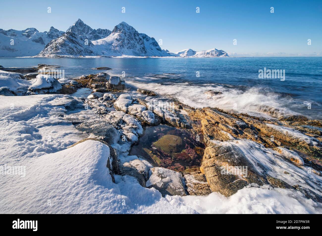 Schneebedeckter Felsstrand mit Gezeitenbecken, Küste mit blauem Meer, hinter Winterbergen, Nordland, Lofoten, Norwegen Stockfoto