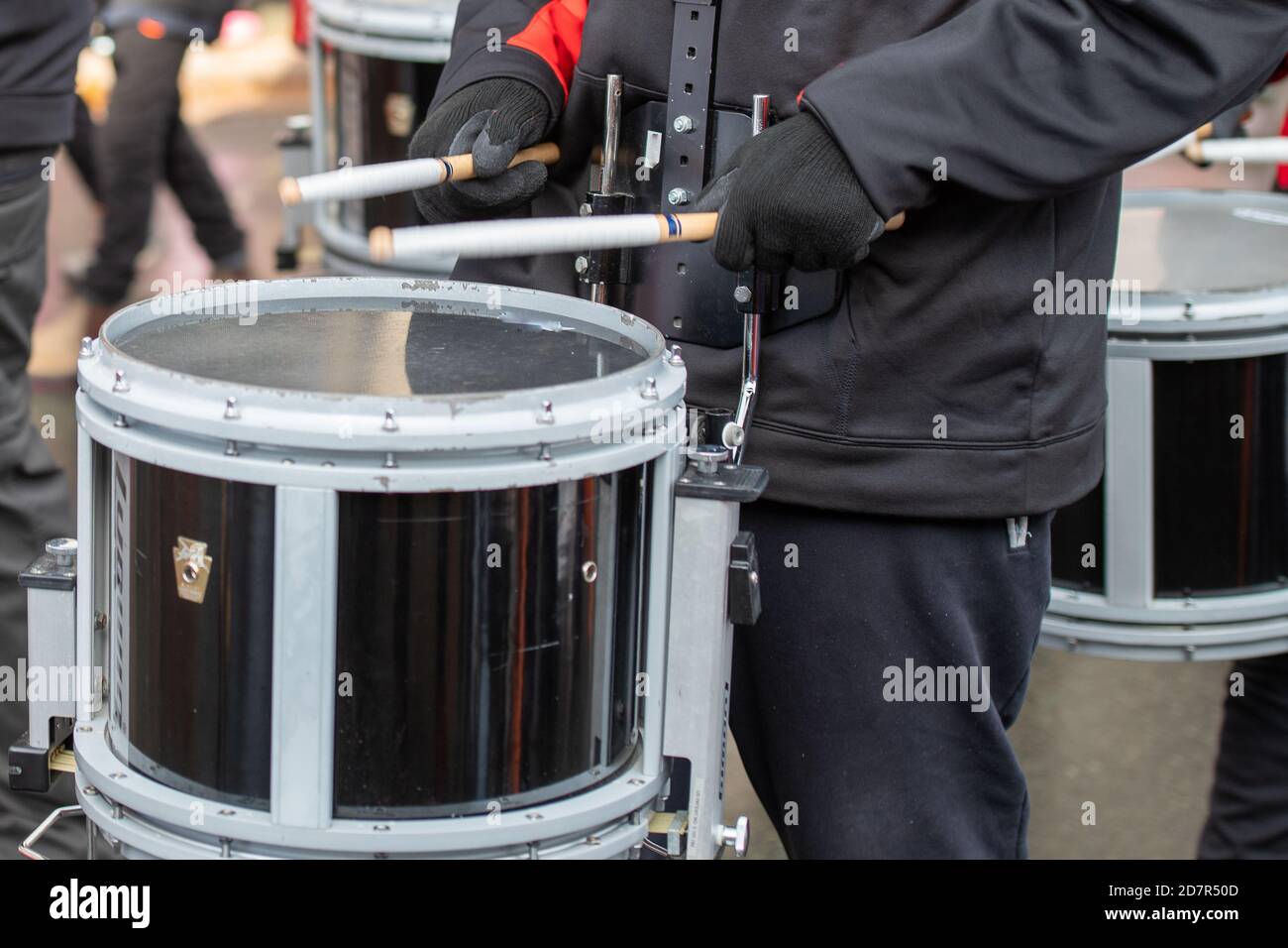 Ein junger Mann in dunkler Kleidung hält bei einem Konzert oder einer Parade im Freien zwei Drumsticks und Beats auf einer schwarz-weißen Trommel. Stockfoto