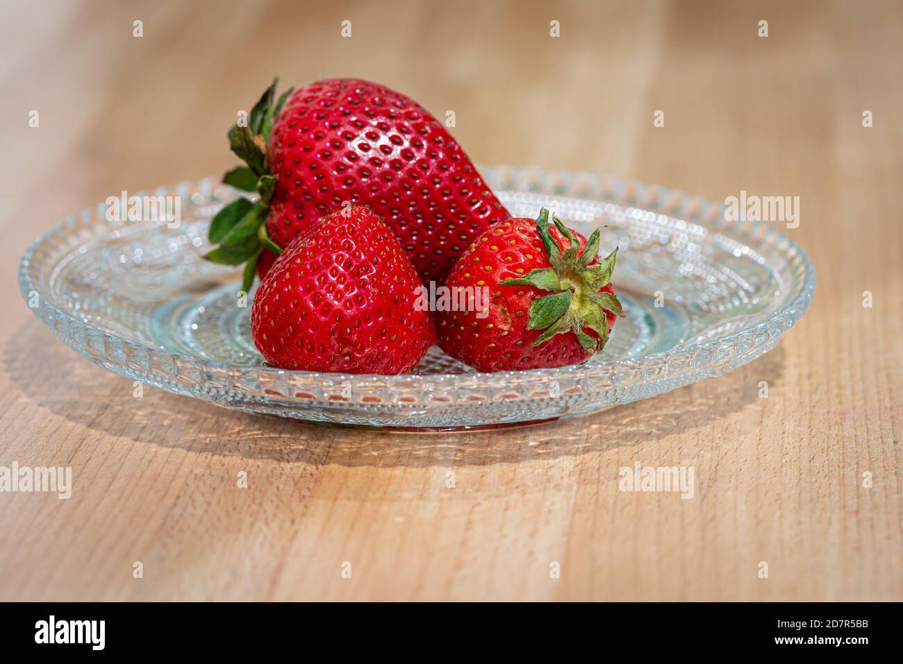 Frische rote Erdbeeren auf einer klaren dekorativen Glasplatte. Der runde Teller sitzt auf einem Holztisch aus Eichenholz. Stockfoto