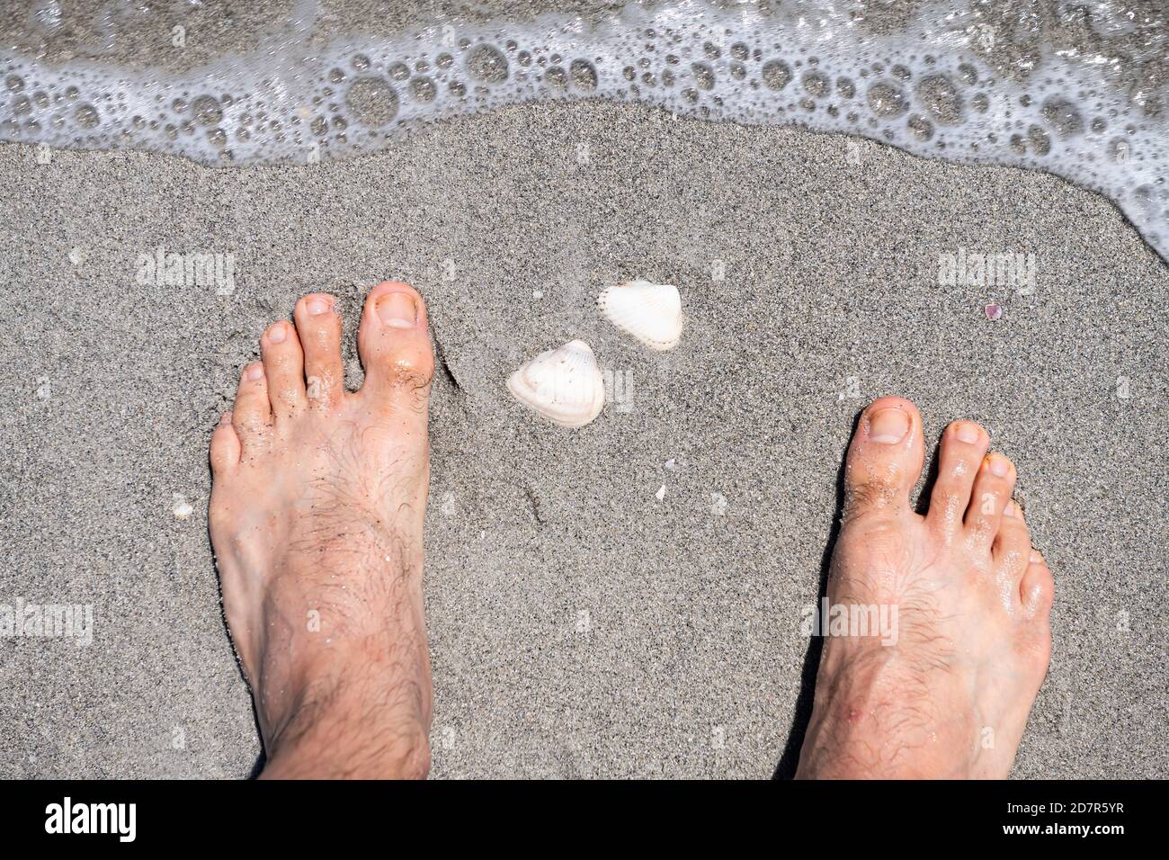 Männliche Füße aus der Nähe Blickpunkt steht bei weißen Muscheln Muscheln während Beschuss Spaß Aktivität auf Sanibel Island, Florida USA auf Ufer Wellenwasser Stockfoto