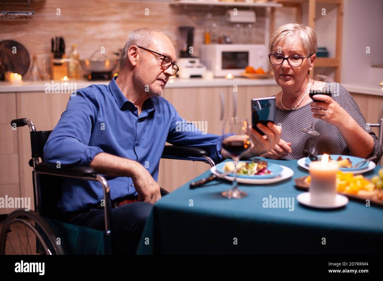 Behinderter Mann mit Smartphone in der Küche beim Essen mit reifen Frau. Bildlauf mit Fotos. Imobilisierter behinderter älterer Ehemann, der am Telefon das festliche Essen genießt. Stockfoto