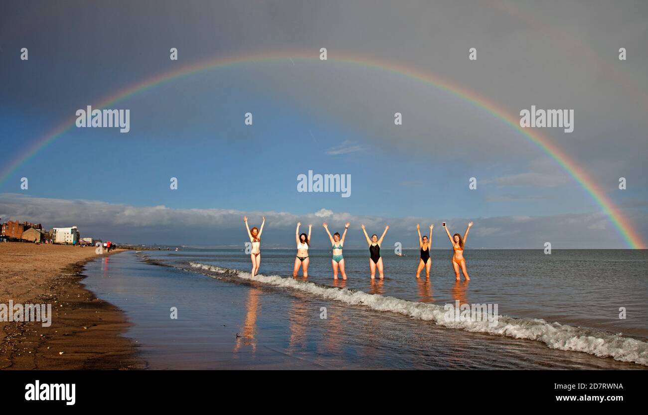 Portobello Beach, Edinburgh, Schottland, Großbritannien. 25. Oktober 2020. Eine Gruppe von sozial distanzierten Studenten, die an der Universität Edinburgh teilnehmen, greifen nach dem Regenbogen, um bessere Dinge zu versprechen, während andere die hellen Bedingungen genießen, bevor der Regen einige Minuten später einströmte. Stockfoto
