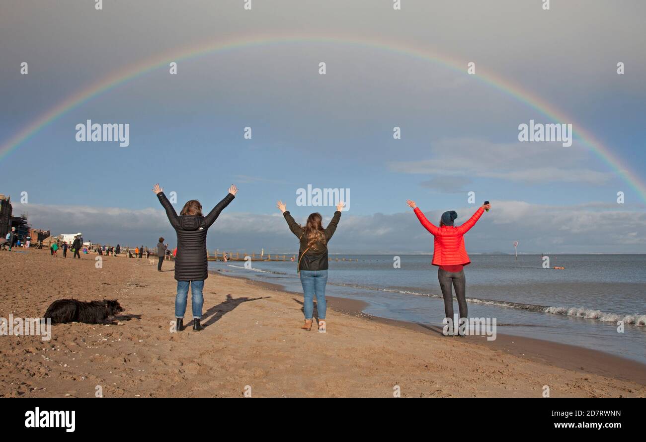 Portobello Beach, Edinburgh, Schottland, Großbritannien. 25. Oktober 2020. Eine Gruppe von sozial distanzierten Freunden greifen nach dem Regenbogen, um bessere Dinge zu versprechen, die kommen, während sie die hellen Bedingungen genießen, bevor der Regen einige Minuten später einströmte. Stockfoto