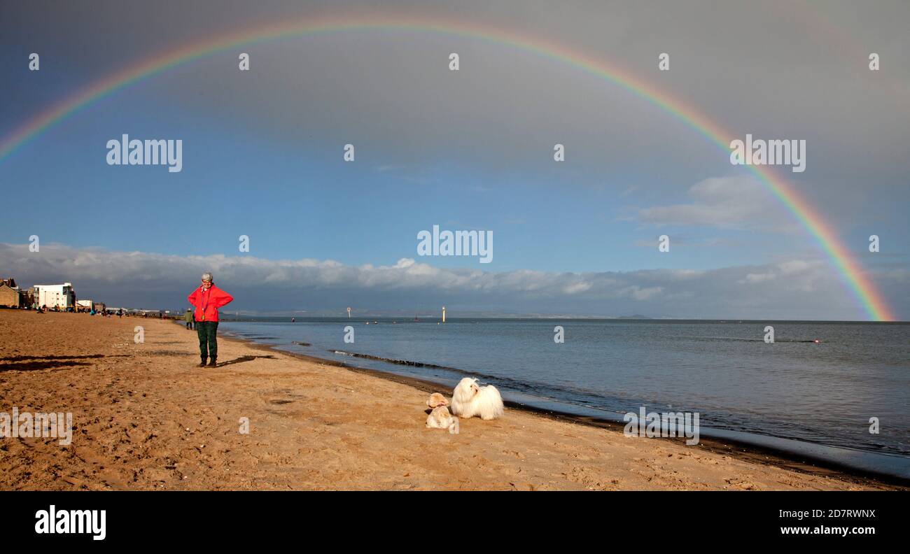 Portobello Beach, Edinburgh, Schottland, Großbritannien. 25. Oktober 2020. Eine Dame wartet auf ihren Hund, als ein Regenbogen über dem Sandstrand und Firth of Forth erscheint, während sie die hellen Bedingungen genießt, bevor der Regen einige Minuten später einströmte. Stockfoto