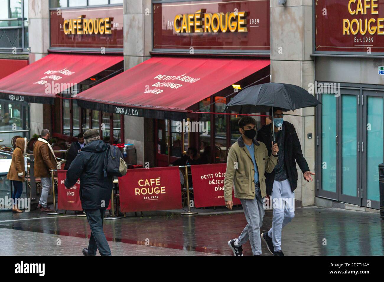 Männer laufen in Birmingham mit einem Regenschirm tragen Gesichtsmasken, wie die Stadt in Coronavirus Alarm Tier 2, High Alert - Cafe Rouge im Hintergrund Stockfoto