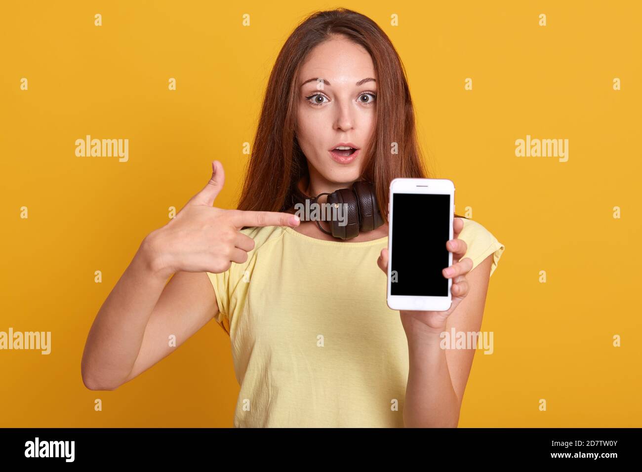 Studio sho von liebenswert Frau zeigt Telefon mit leerem Bildschirm und zeigt auf sie mit ihrem Zeigefinger, hat Gesichtsausdruck erstaunt, hält Mund o Stockfoto