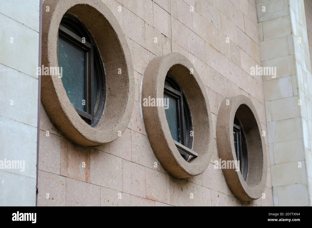 Drei runde Fenster mit quadratischen Fensterrahmen innen an der Fassade der Steinmauer. Hohe Gebäudefassade mit einer Reihe von Fenstern. Zeitgenössische Architektur Stockfoto