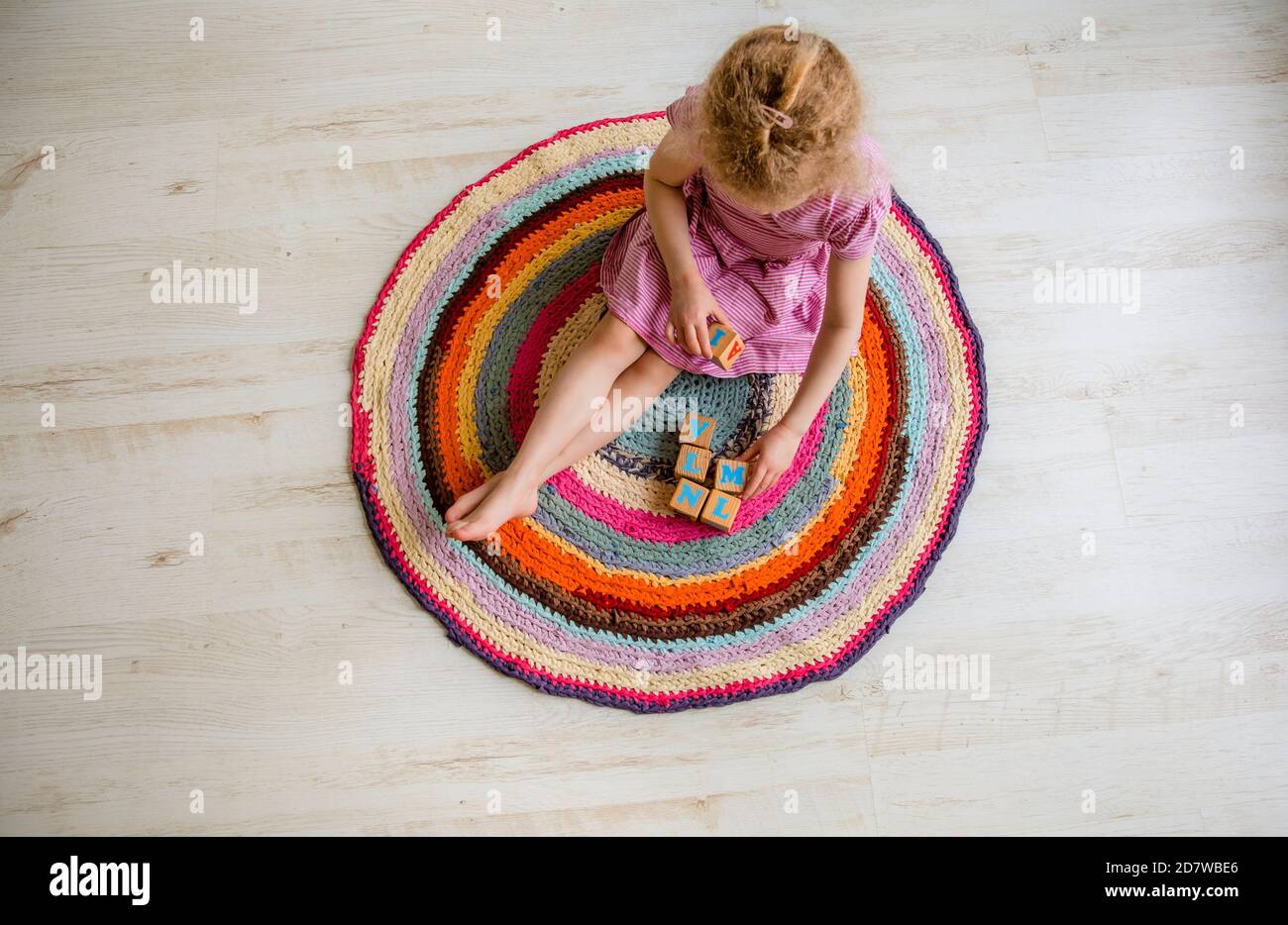 4 Jahre altes kleines Kind sitzt auf runden bunten Häkeln Teppich aus umfunktionierten T-Shirts Garn auf weißem Laminatboden und Spielen Sie natürliche Holzwürfel mit Let Stockfoto
