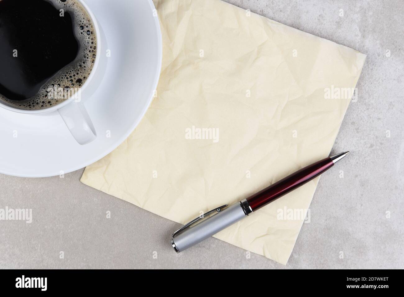 Eine Tasse Kaffee mit einer zerknitterten Serviette und einem Stift auf einem hellgrauen Tisch. Stockfoto
