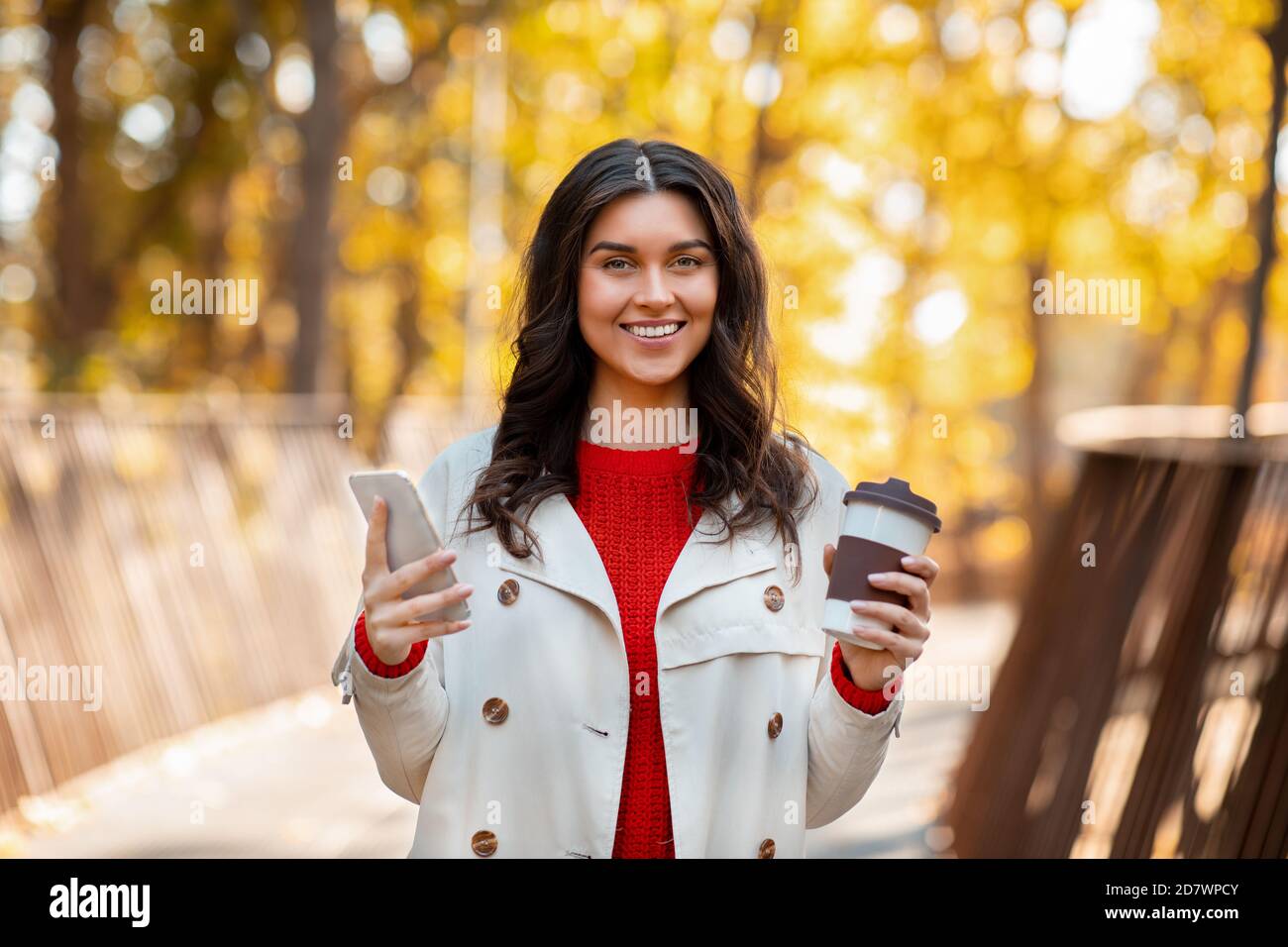 Portrait von hübschen Dame mit Kaffee zu gehen und Smartphone Lächelnd und Blick auf die Kamera im Freien an einem kühlen Herbsttag Stockfoto