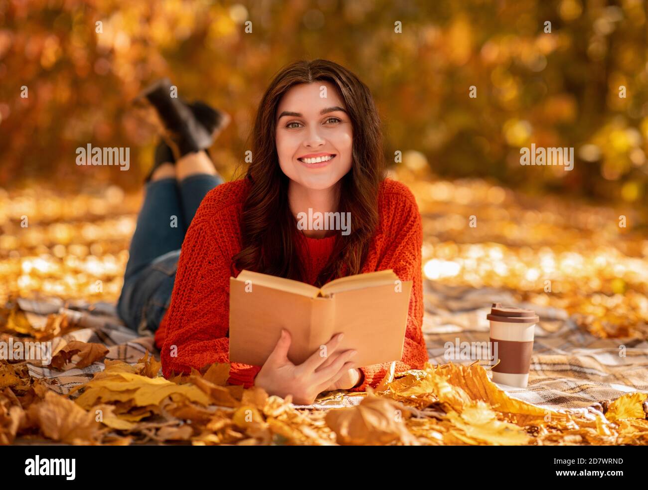 Lächelnde junge Frau mit Buch und Kaffee zum liegen gehen Auf Decke zwischen herbstlichen Blättern und warmen Herbst genießen Tag Stockfoto