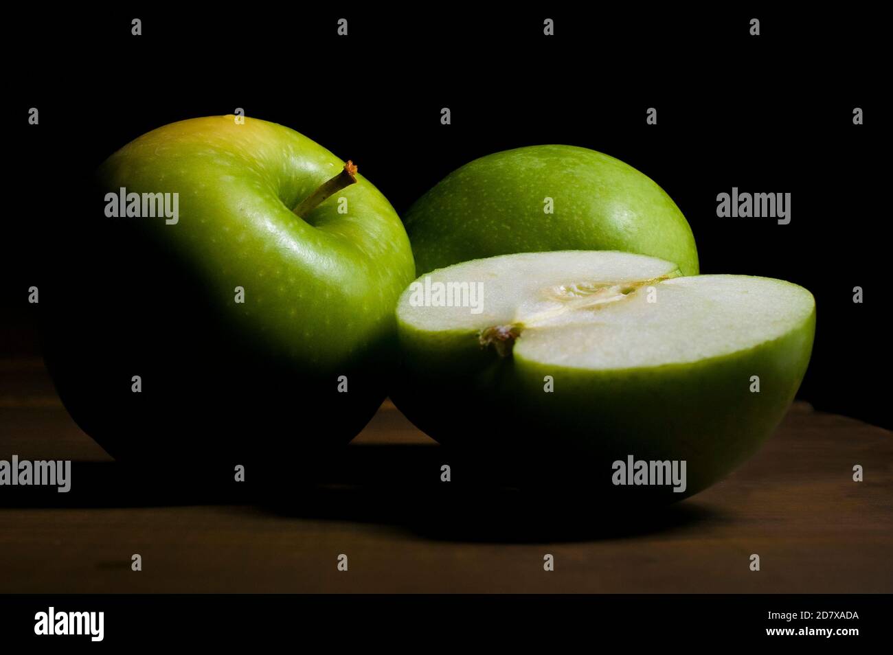 Stillleben, Grüne Apfelhälfte und grüne Äpfel mit einem Chiaroscuro, Barockstil mit schwarzem Hintergrund und auf einem rustikalen Holztisch Stockfoto