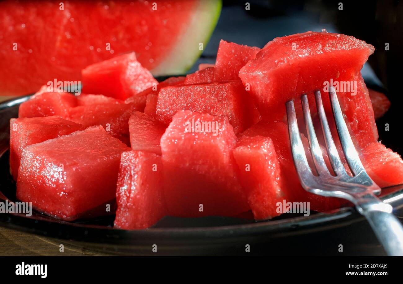 Eine Gabel mit einem Würfel Wassermelone über einige mehr Wassermelonenwürfel auf einem schwarzen Teller und eine Wassermelonenscheibe Und eine ganze Wassermelone im Hintergrund Stockfoto