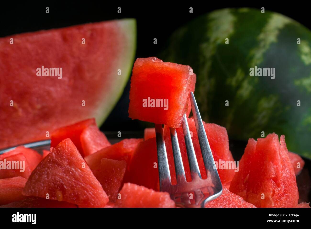 Eine Gabel mit einem Würfel Wassermelone über einige mehr Wassermelonenwürfel auf einem schwarzen Teller und eine Wassermelonenscheibe Und eine ganze Wassermelone im Hintergrund Stockfoto